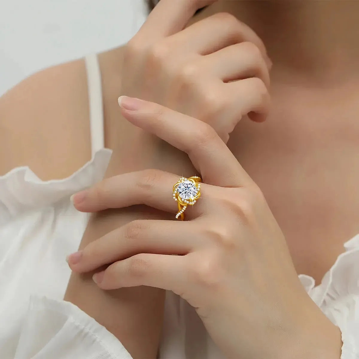 A person wearing a gold ring with a large, round gemstone on their finger. The person is dressed in a white, ruffled top and is gently touching their chin with their other hand.