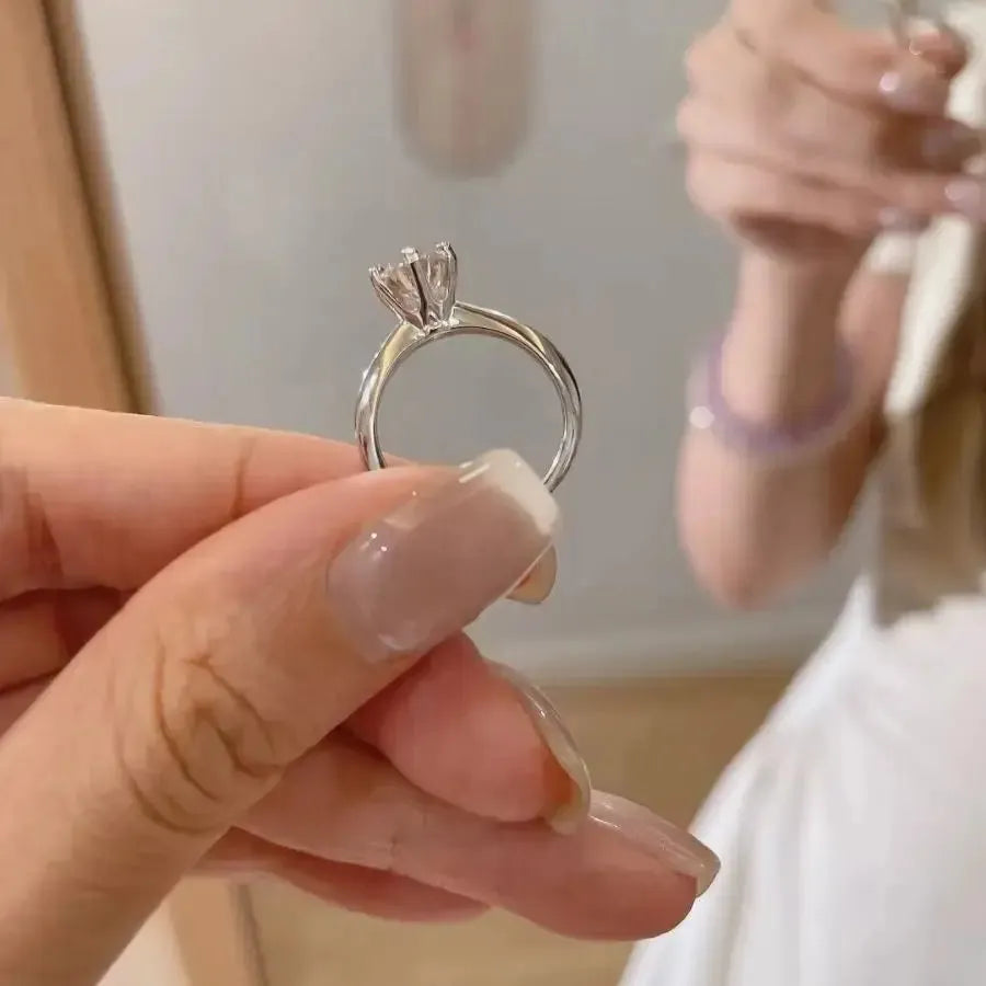 A person holding a silver ring with a solitaire diamond setting, with another person in the background wearing a white outfit and a bracelet.