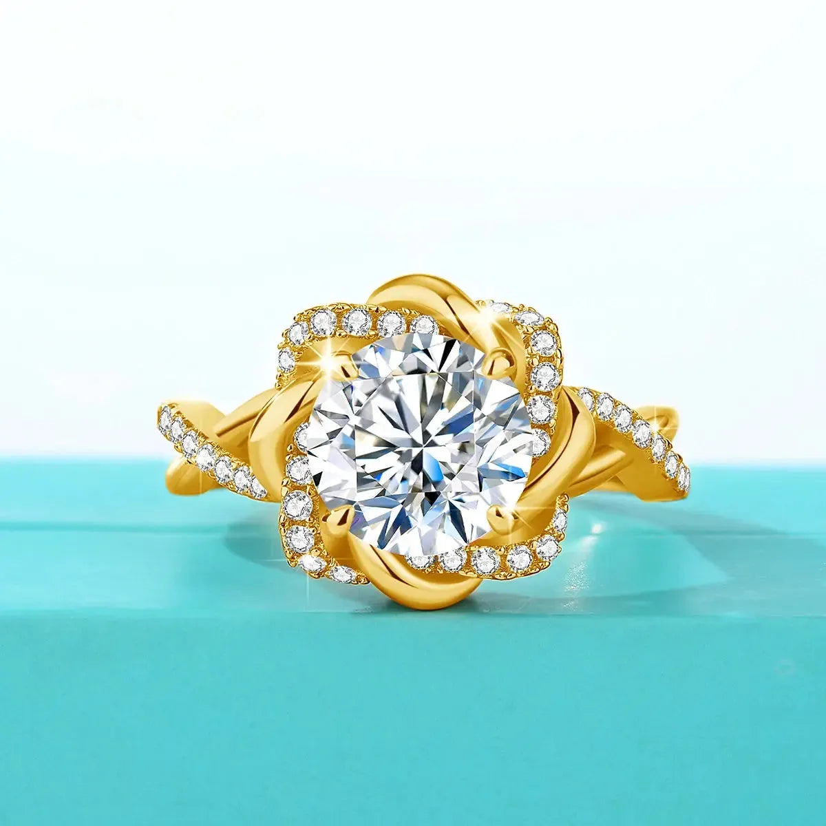 A gold ring with a large round diamond in the center, surrounded by smaller diamonds in a floral design, placed on a light blue surface.