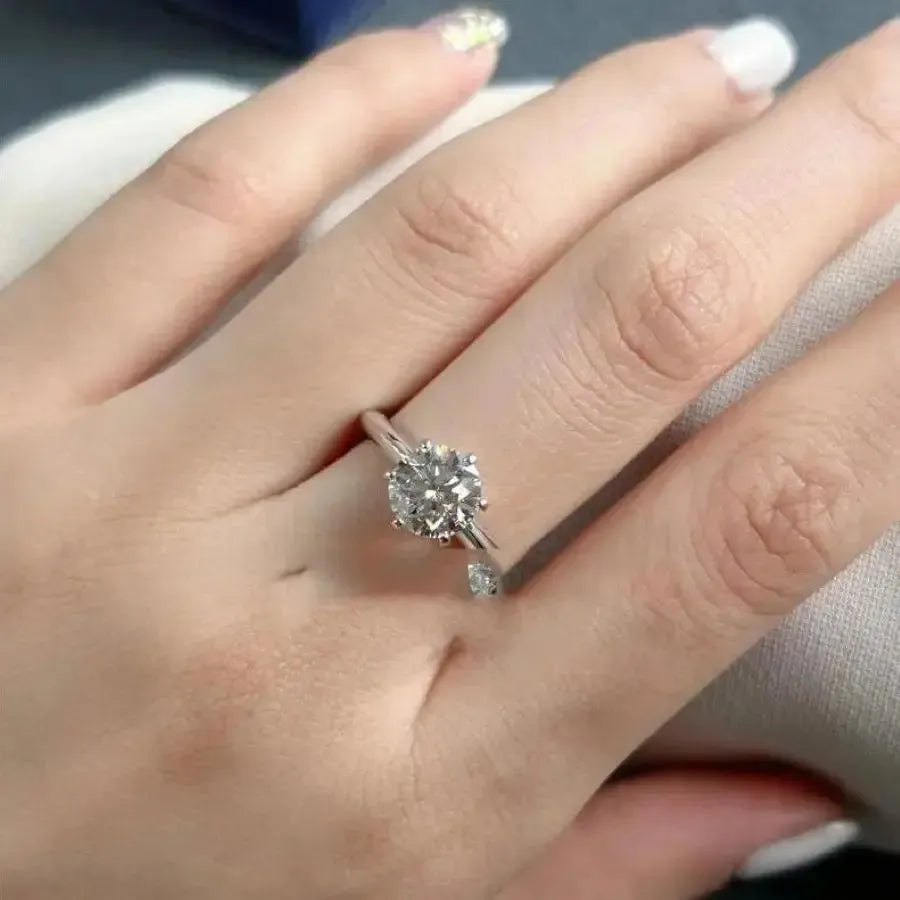 A hand wearing a silver ring with a large, round diamond in the center. The nails are manicured with a light color and one nail has a decorative design.